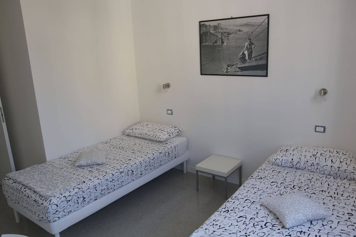A room with two single beds, each with a pillow and a small bedside table. A framed picture hangs on the wall above the beds.