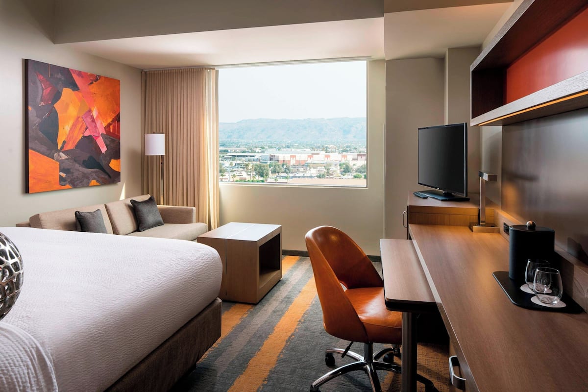 Habitación de hotel con cama, escritorio, silla, televisor y ventana grande con vista a la ciudad.