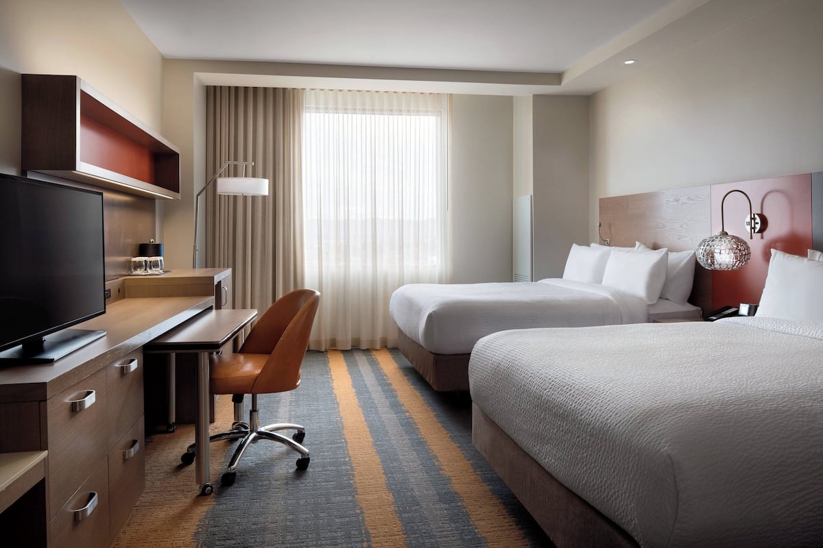 Habitación de hotel con escritorio, silla, dos camas, televisor y un amplio ventanal con cortinas.