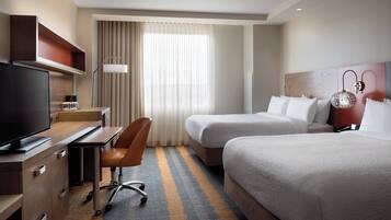 A hotel room with a desk, chair, two beds, a TV, and a large window with curtains.