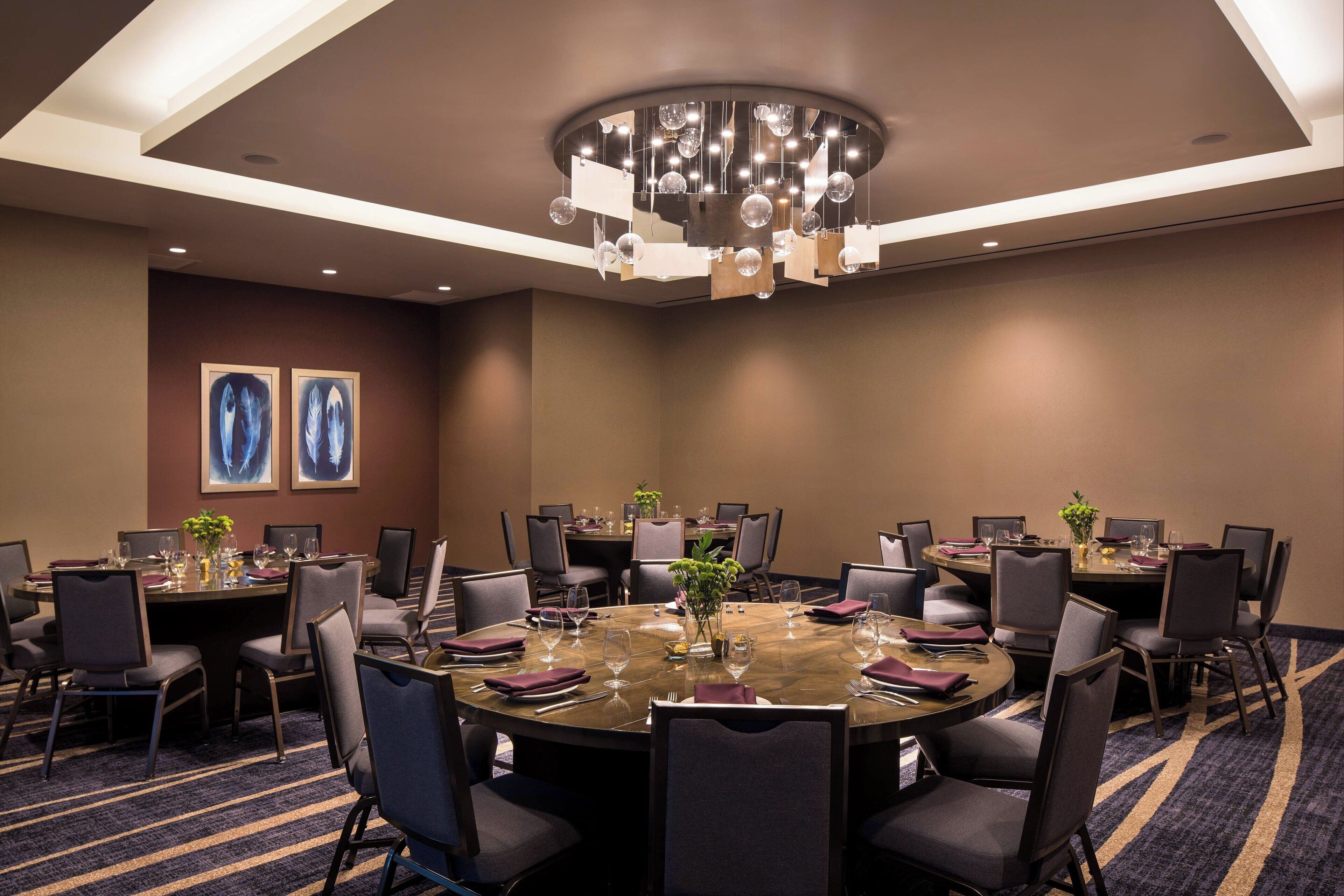 A conference room with a round table, multiple chairs, and a chandelier. There are two framed pictures on the wall.