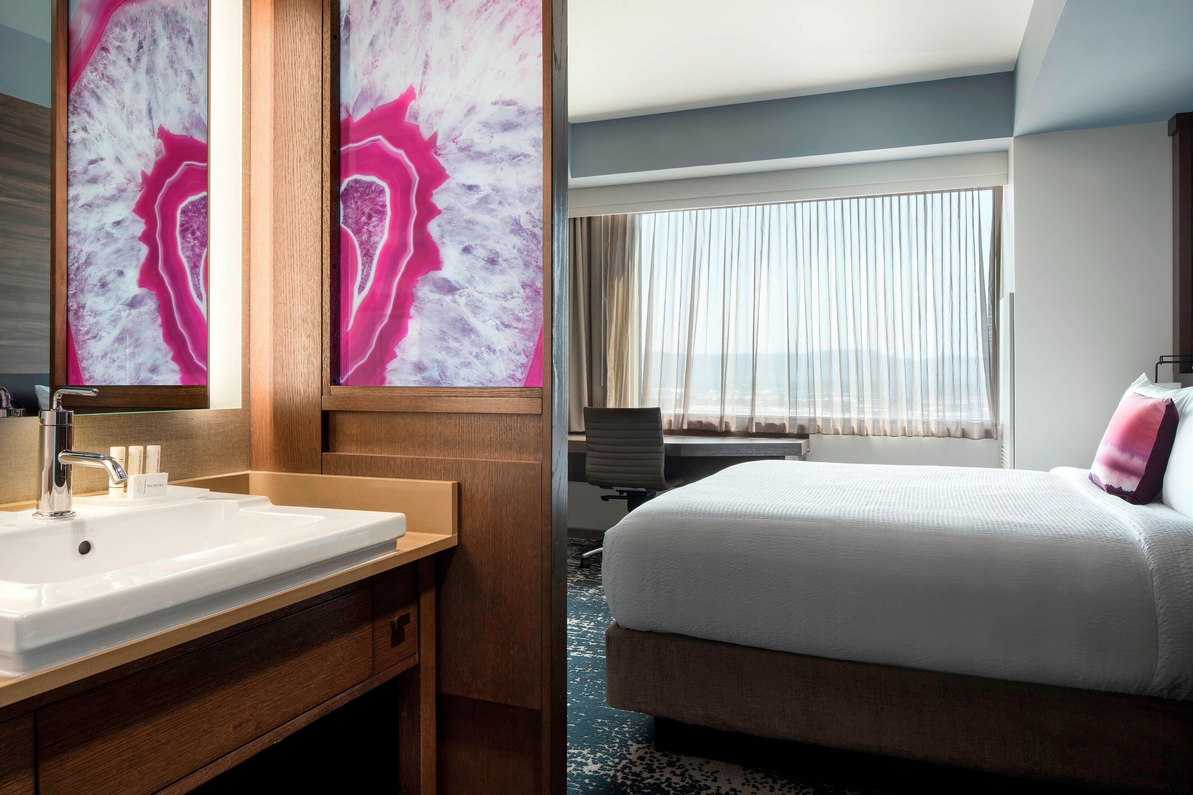 A hotel room with a large bed, a wooden vanity with a sink, and a large mirror. There is a window with curtains and a view of the city.