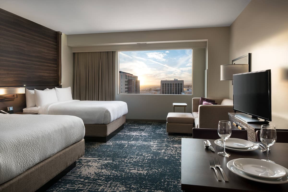 A hotel room with a large bed, a desk with dining set, a TV, and a view of the cityscape.