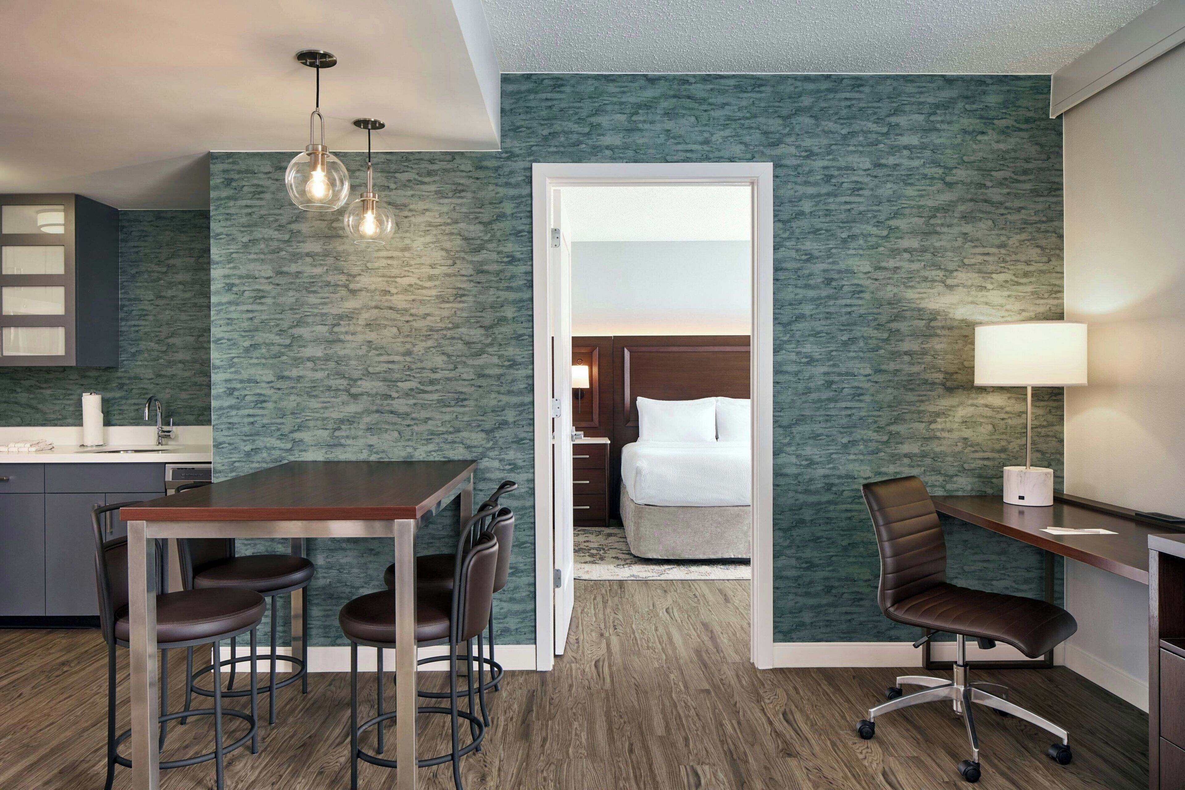 A hotel room with a kitchenette, a dining area with stools, a bed, a desk with a chair, and a nightstand with a lamp.