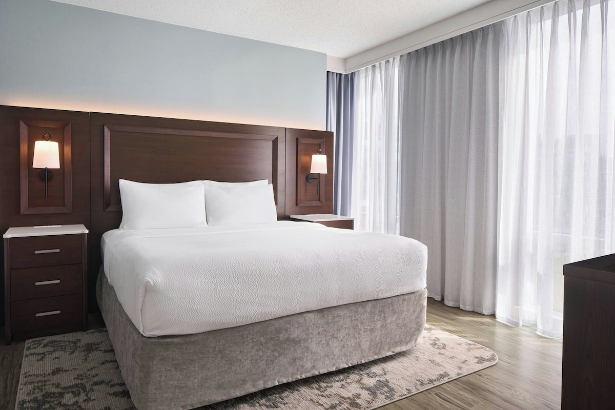 Ein Hotelzimmer mit einem großen Bett, zwei Nachttischen mit Lampen, einem Holz-Kopfteil und einem großen Fenster mit durchsichtigen Vorhängen.