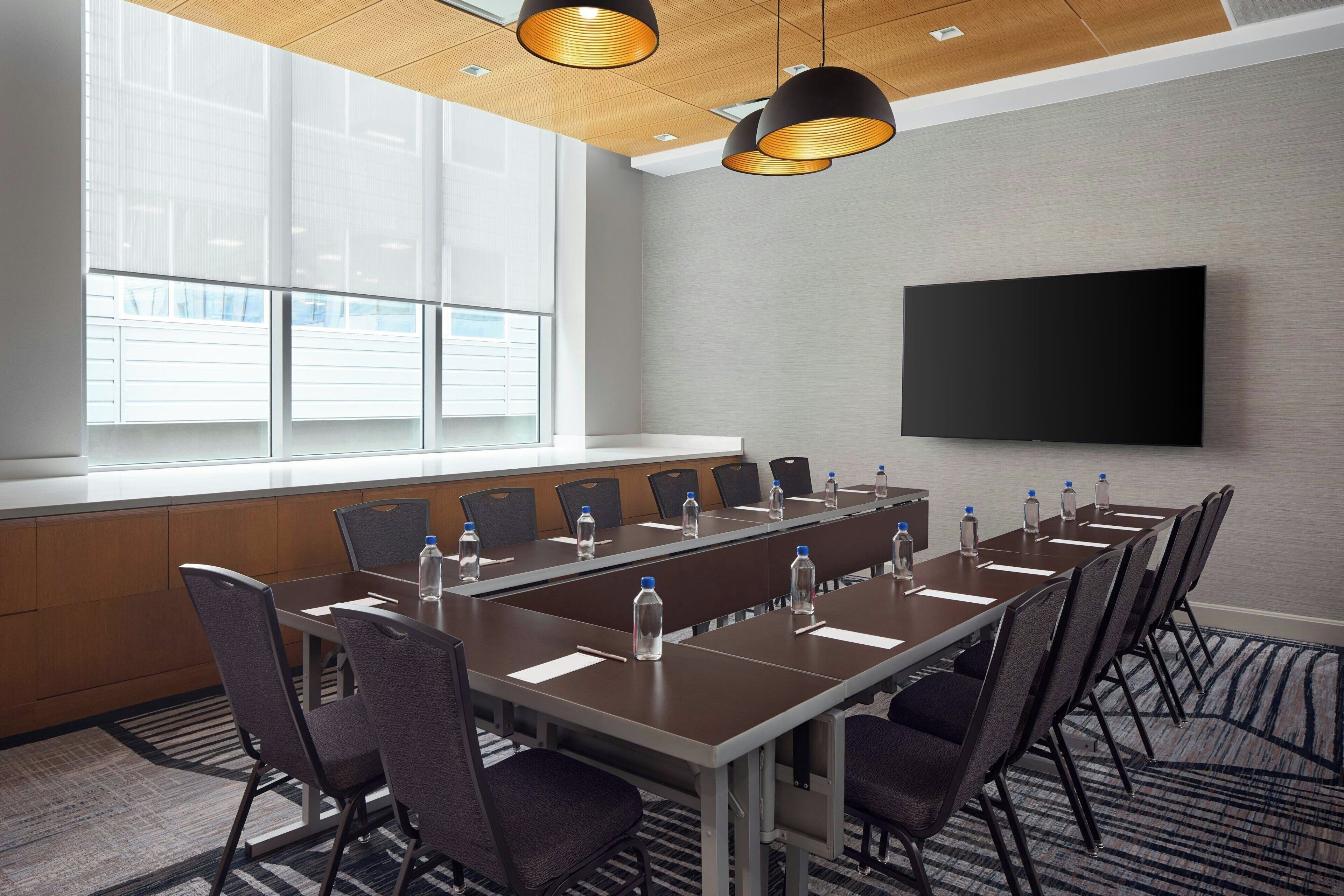 A conference room with a long table, chairs, water bottles, a flat-screen TV, and large windows.