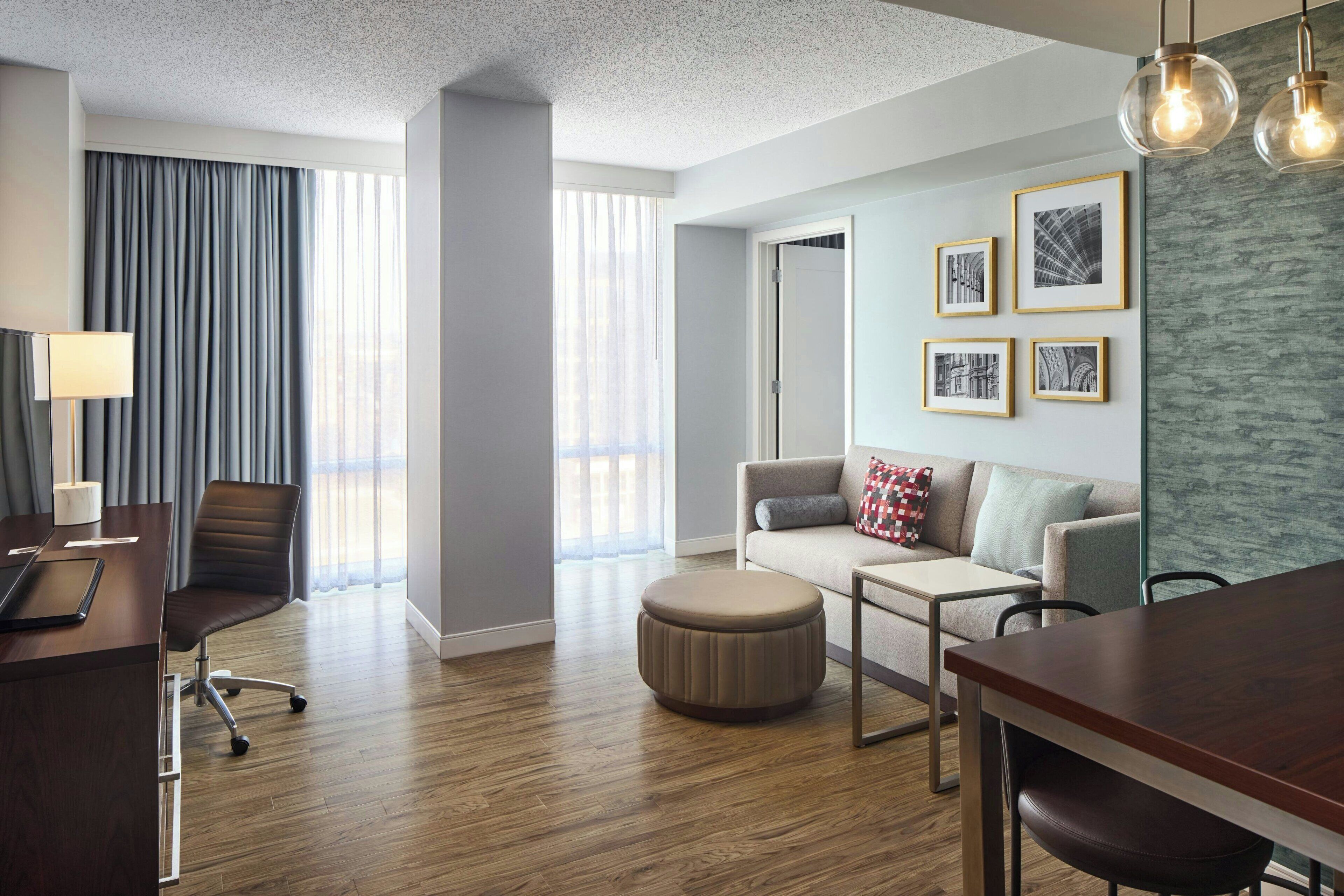A modern hotel room with a wooden floor, a desk with a lamp, a sofa, a coffee table, and framed pictures on the wall.