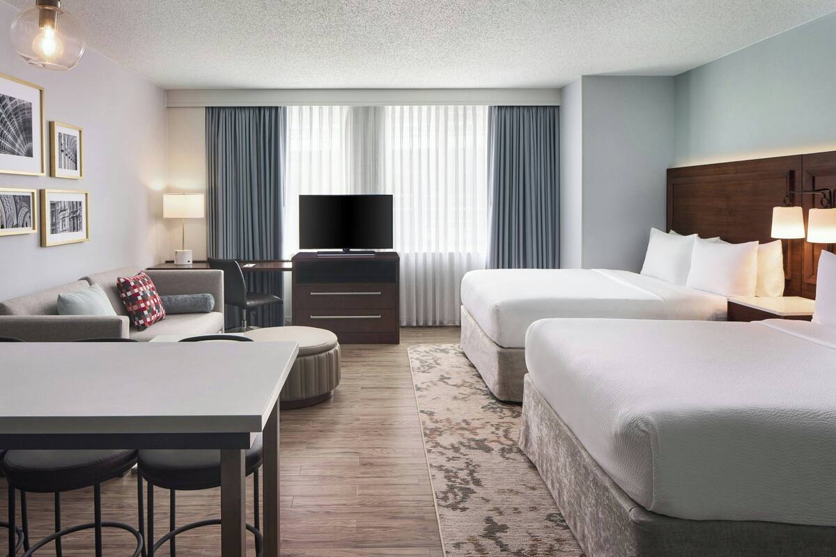 Ein Hotelzimmer mit einem Bett, einem Sofa, einem Fernseher, einem Schreibtisch mit Stühlen und einem großen Fenster mit Vorhängen.