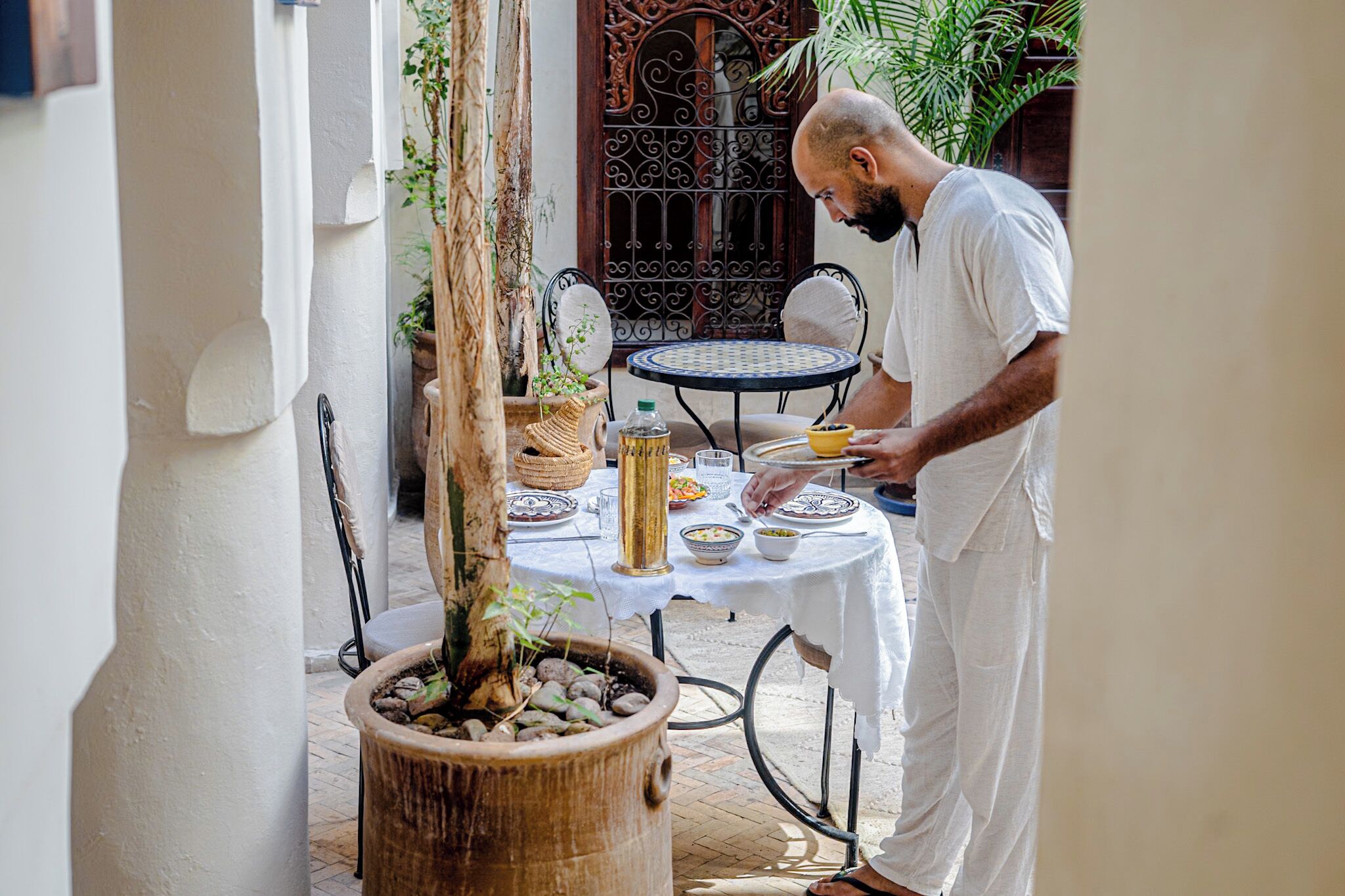 Um homem servindo comida em uma mesa externa com toalha branca, rodeado por plantas em vasos e uma porta decorativa ao fundo.