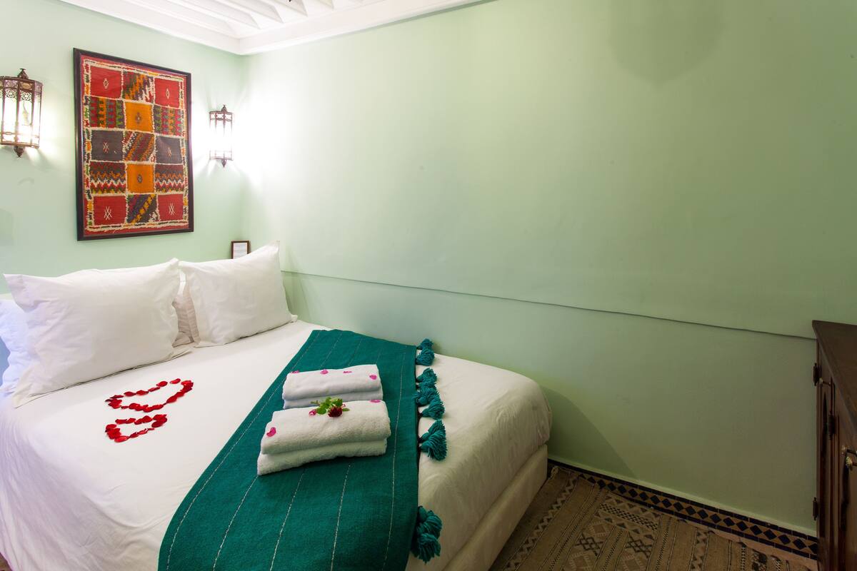 A neatly made bed with white linens, a teal throw blanket, and a heart-shaped decoration on the pillow.