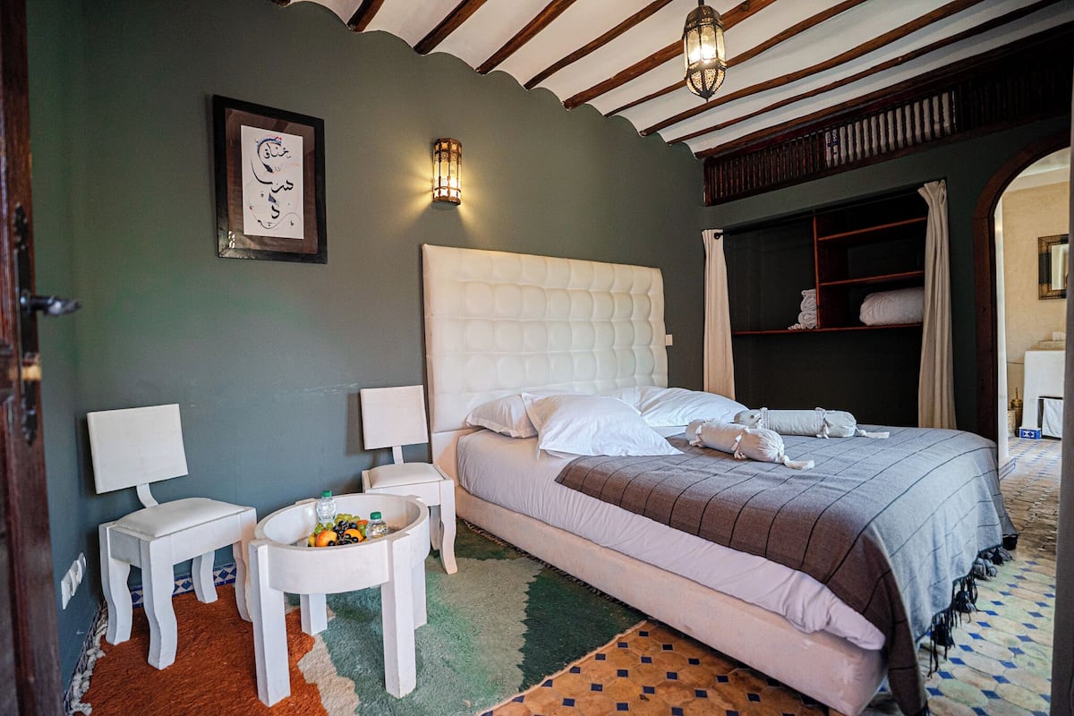 A bedroom with a large bed, a small table with a bottle and a bowl, a chair, and a framed picture on the wall.