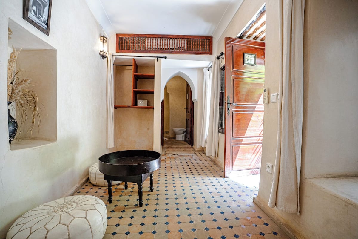 A narrow hallway with a tiled floor, a black basin on a stool, and a wooden door with a glass panel.