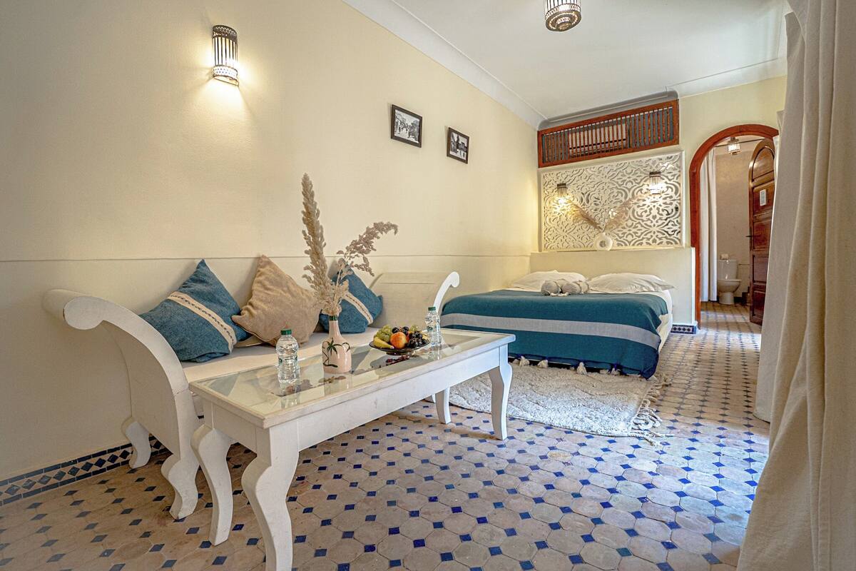 A hotel room with a sofa, a glass coffee table, a bed with a blue and white striped bedspread, and a patterned wall decoration.