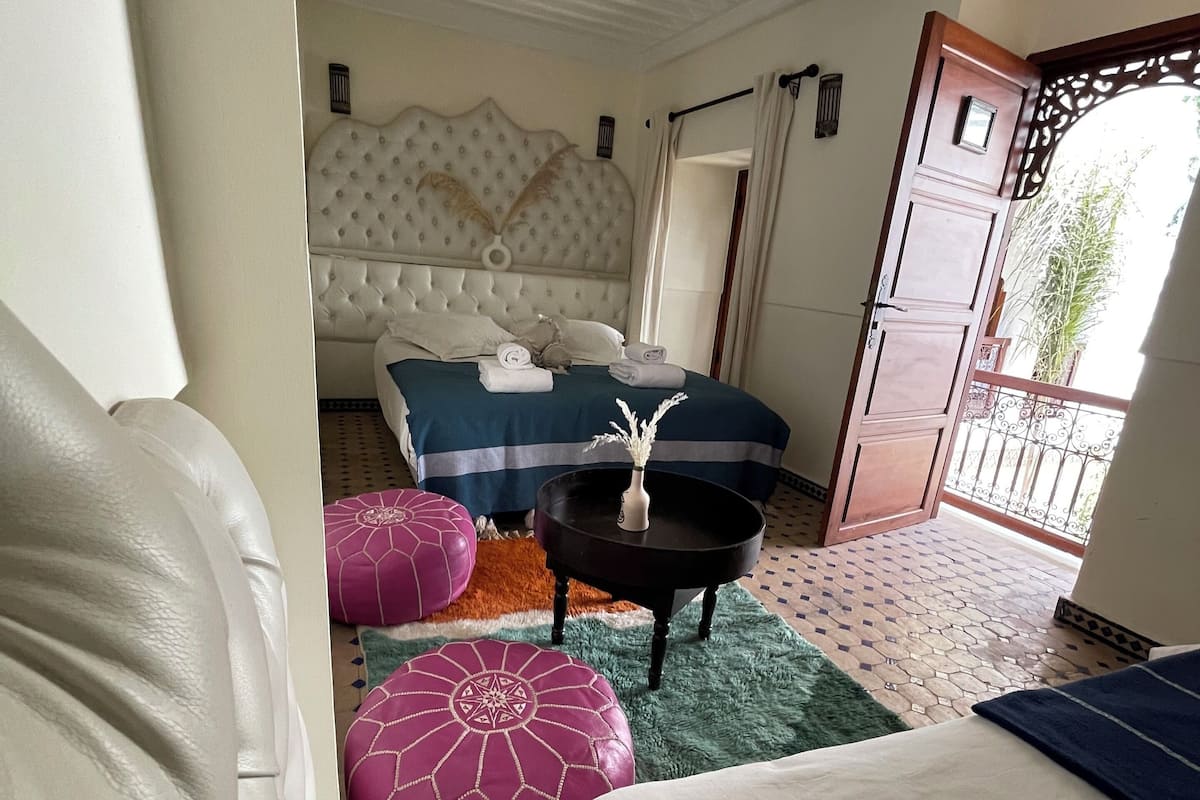 A bedroom with a large bed, a nightstand, a decorative lamp, and a view of the outside.