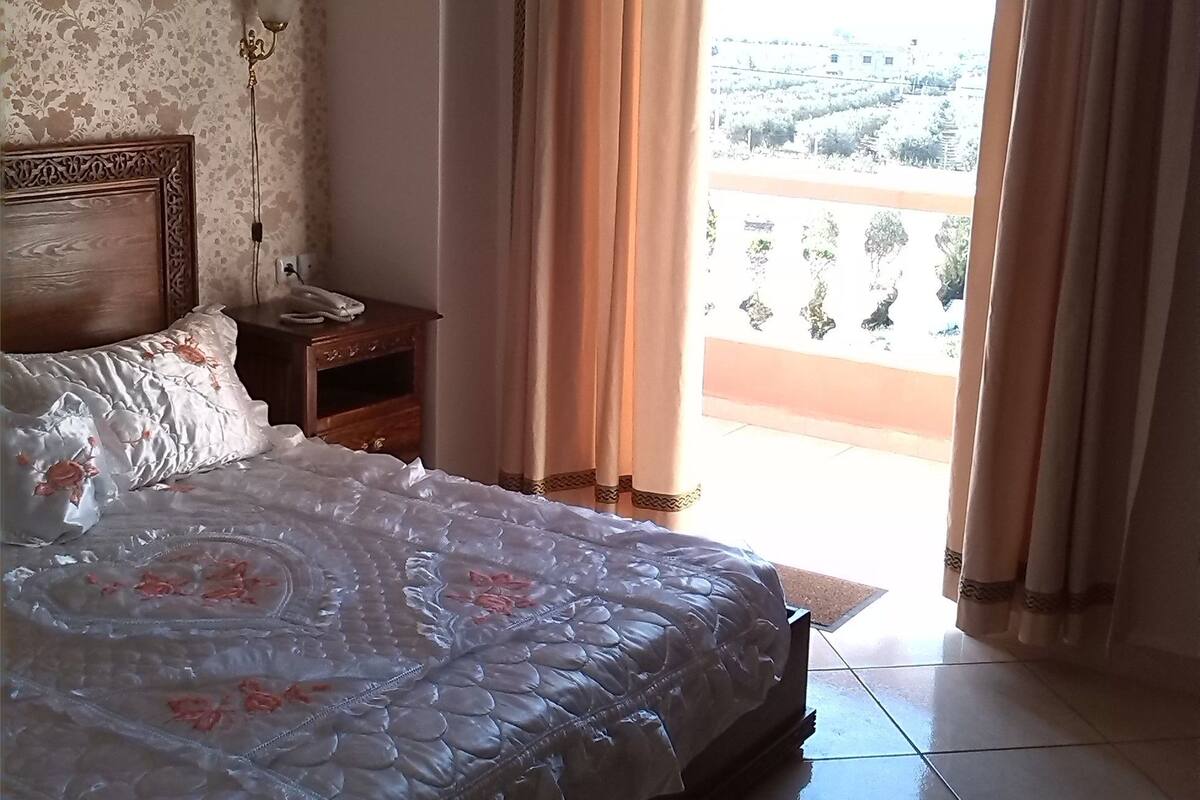 A bedroom with a bed, a nightstand, a telephone, and a window with curtains.