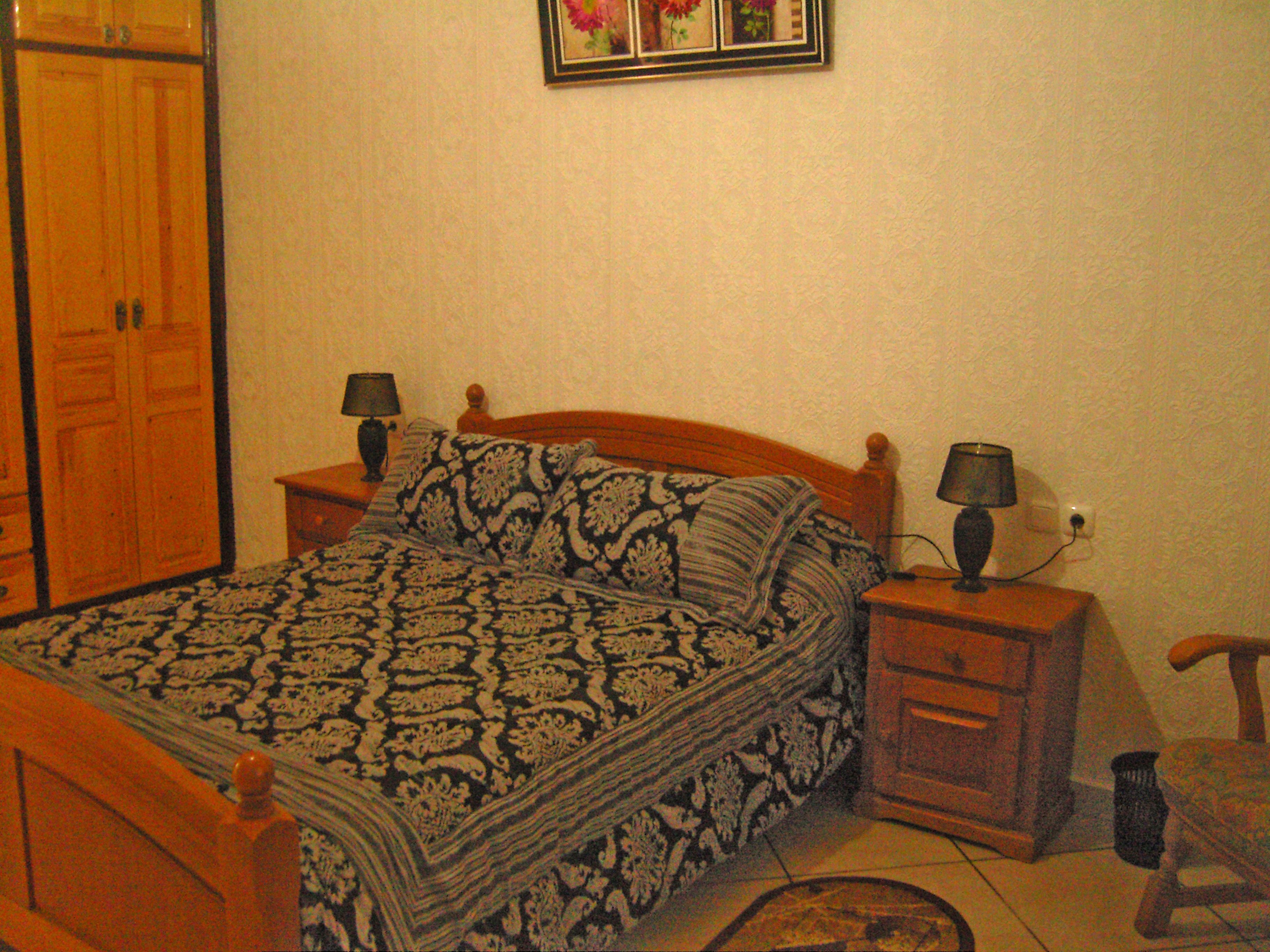 Ein Schlafzimmer mit einem hölzernen Bett, gemusterten Betttextilien, zwei Nachttischen mit Lampen, einem Beistelltisch und einem Sessel.