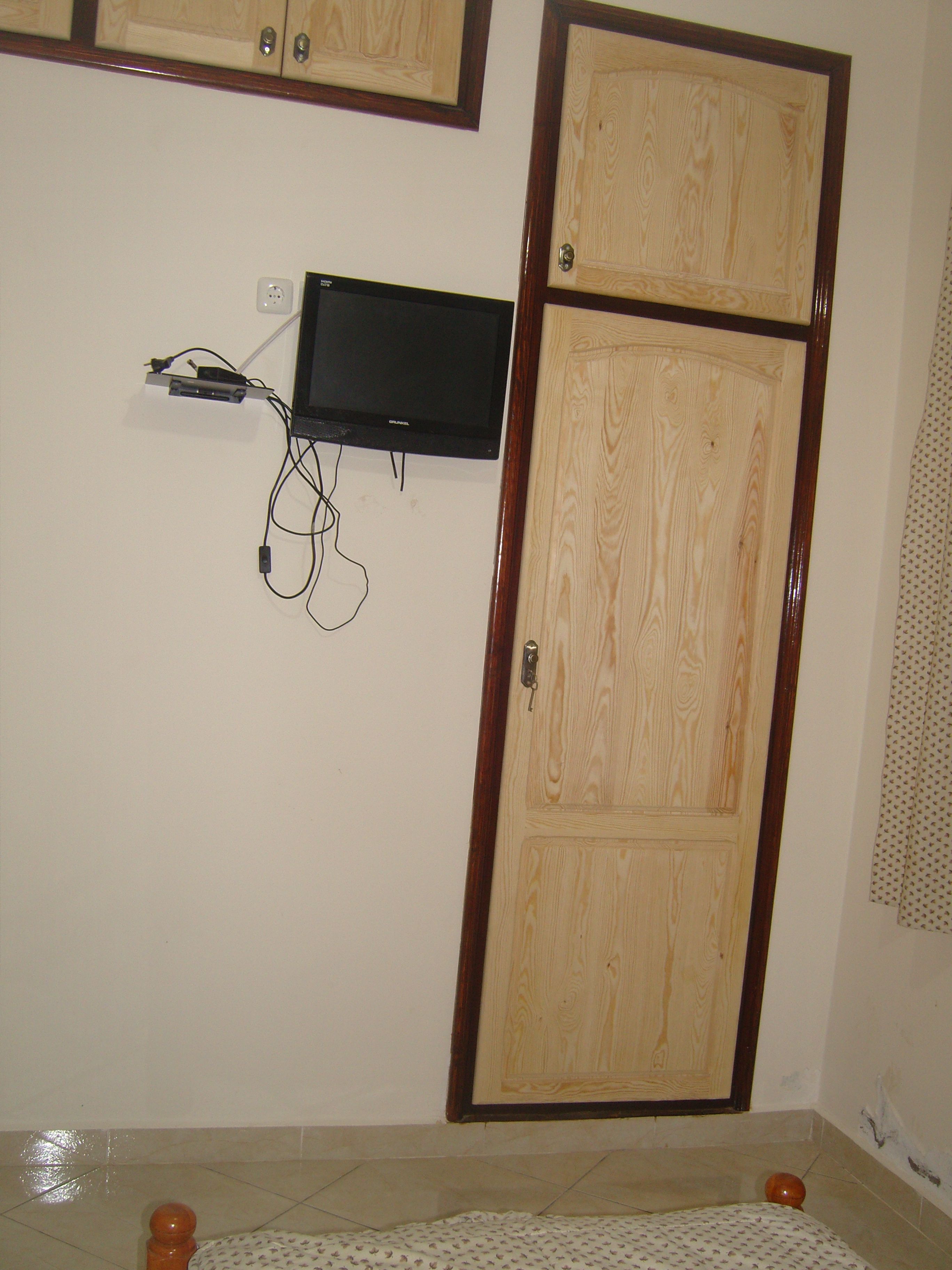 A room with a wooden wardrobe, a mounted television, and a tiled floor.