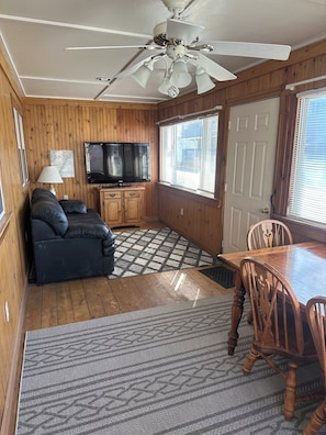 A compact living space with a ceiling fan, a television, a sofa, a dining table with chairs, and wooden walls. - Nautical Beach Properties (Hampton)
