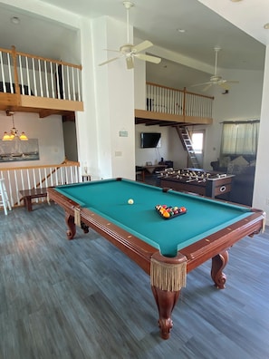 A pool table in a spacious living area with a high ceiling, a staircase, and a television mounted on the wall. - Nautical Beach Properties (Hampton)