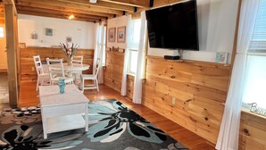 A cozy room with a wooden floor, a round dining table with chairs, a flat-screen TV mounted on the wall, and a window with curtains. - Nautical Beach Properties (Hampton)
