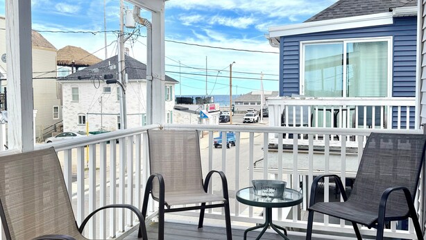 A covered outdoor area with a wooden deck, metal chairs, a glass table, and a view of a building with a large window. - Nautical Beach Properties (Hampton)