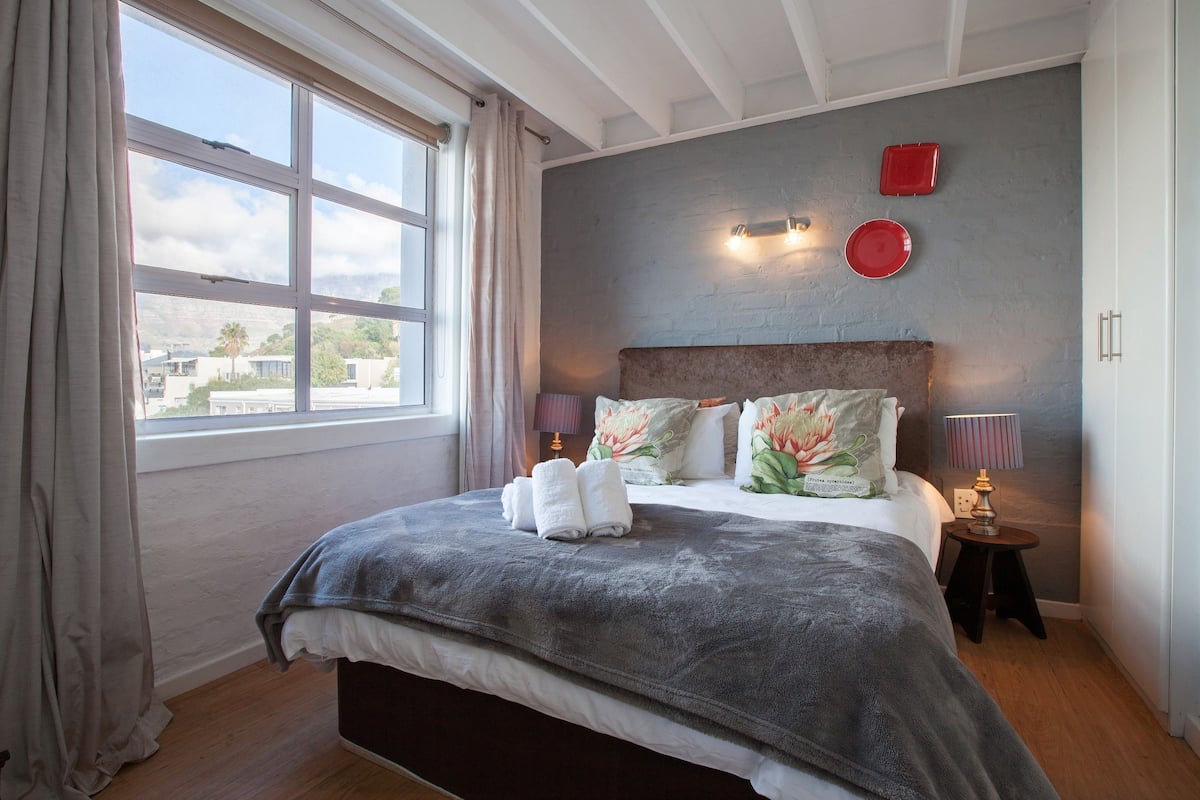 A bedroom with a large window, a bed with a grey blanket, two bedside lamps, and a red plate on the wall.