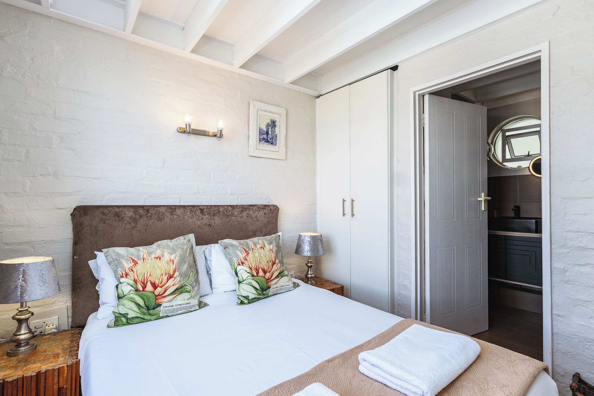 Un dormitorio con cama, lámparas de noche, mesita de noche, una puerta que da al baño y un cuadro colgado en la pared.