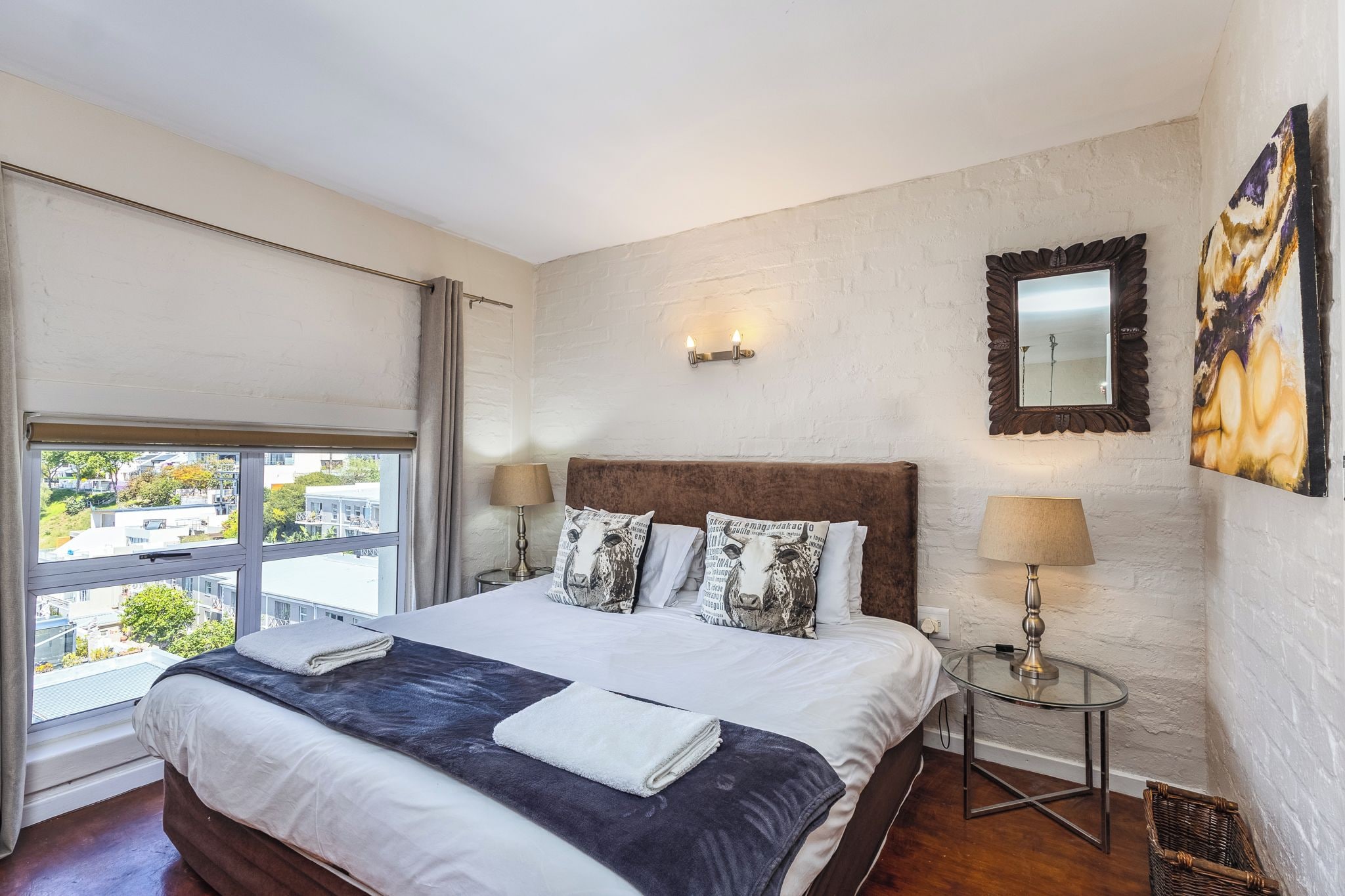 A hotel room with a large bed, two bedside lamps, a window with curtains, a wall-mounted mirror, and a painting.