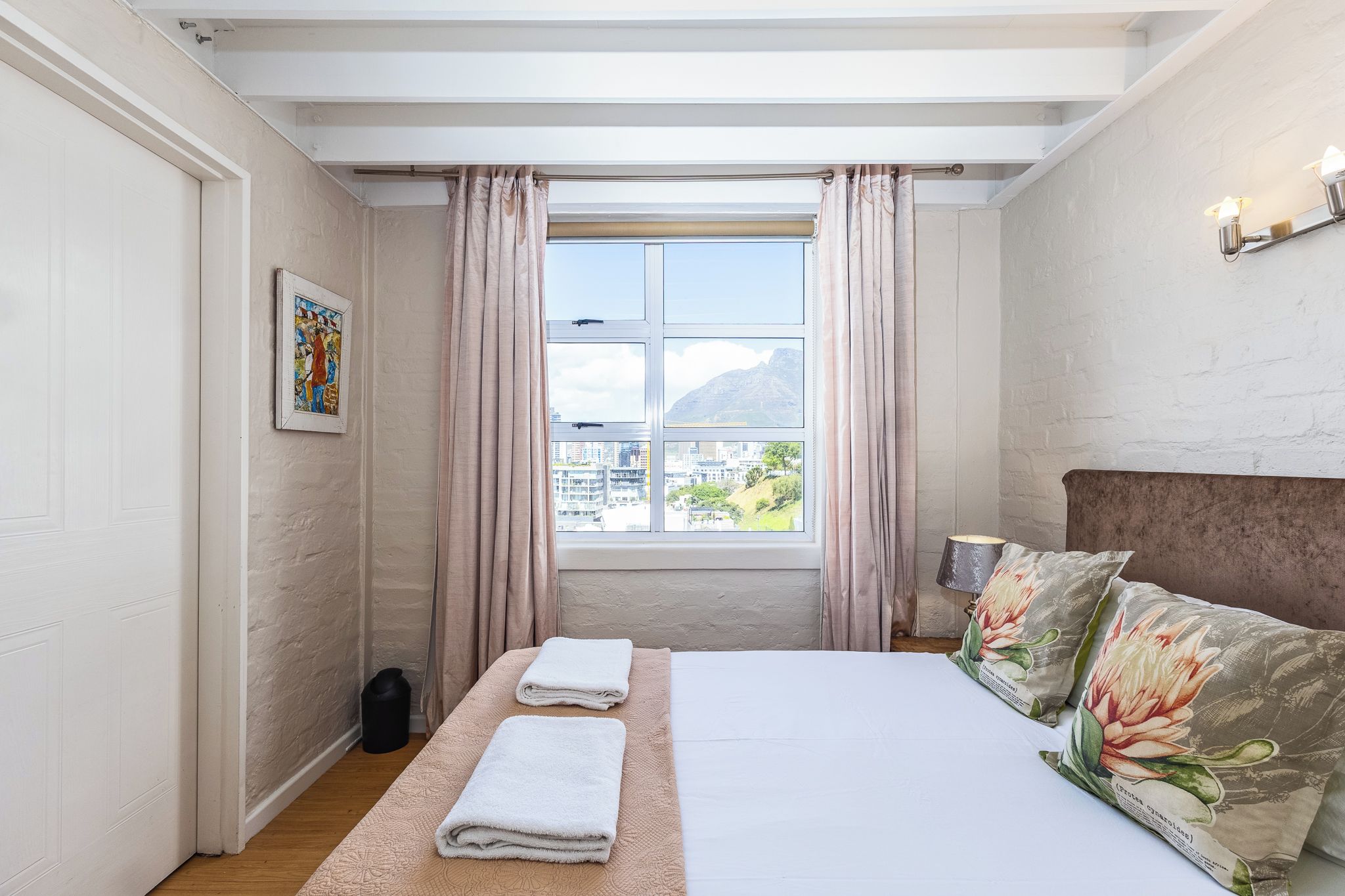 A bedroom with a bed, two towels, a window with curtains, a picture on the wall, and a lamp.