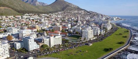 Un paisaje urbano costero con una montaña destacada al fondo, un paseo marítimo en la playa y edificios urbanos densos.