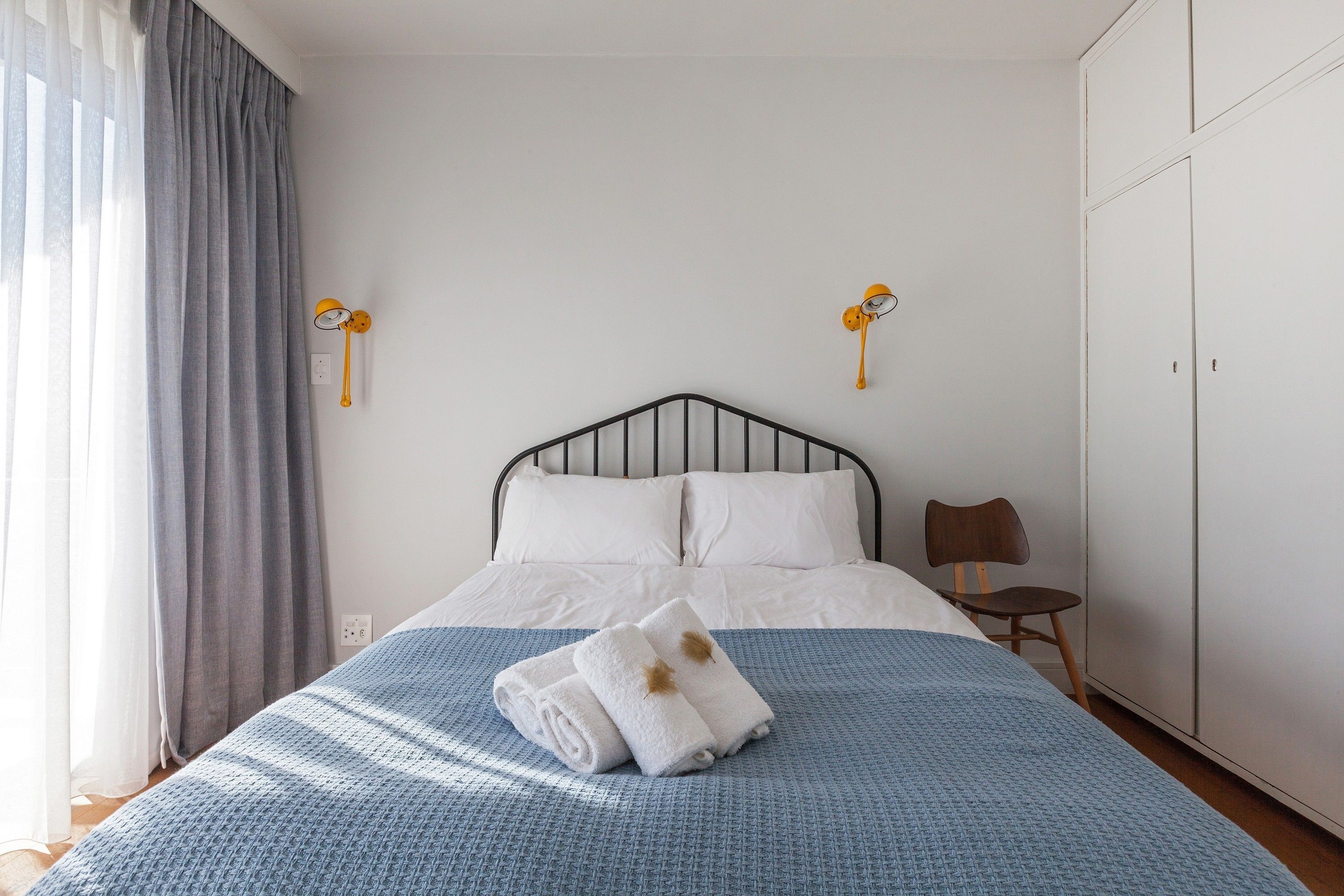 A neatly made bed with white linens and a blue blanket, a wooden chair, and a wardrobe in a simple, clean room.