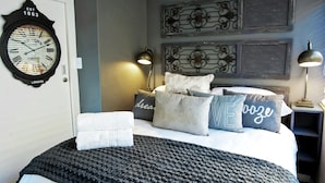 Un lit bien fait, recouvert d’une couverture tricotée de couleur sombre, agrémenté de coussins décoratifs et orné d’une horloge murale.