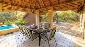 Zona pranzo all'aperto con tetto di paglia, piscina e vista sugli alberi circostanti.