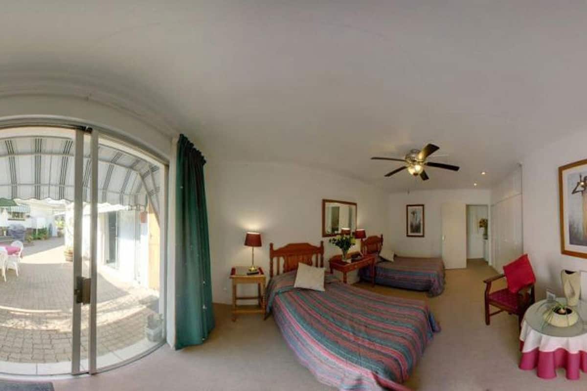 Ein Hotelzimmer mit einem Bett, zwei Nachttischen, einem Sessel, einem kleinen Tisch, einem Deckenventilator, einem Fernseher und einem großen Fenster mit Vorhängen.