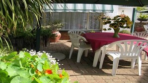 Un espace extérieur aménagé en terrasse, avec une table ronde recouverte d’une nappe rouge, entourée de chaises en plastique blanches. On peut voir, au premier plan, des plantes en pot et diverses fleurs.