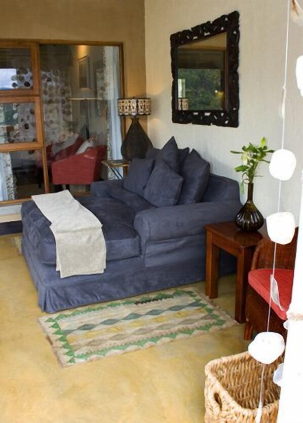 A cozy living room with a grey sofa, a small wooden side table, a floor lamp, and a woven basket. - Tanamera Lodge (Sabie)