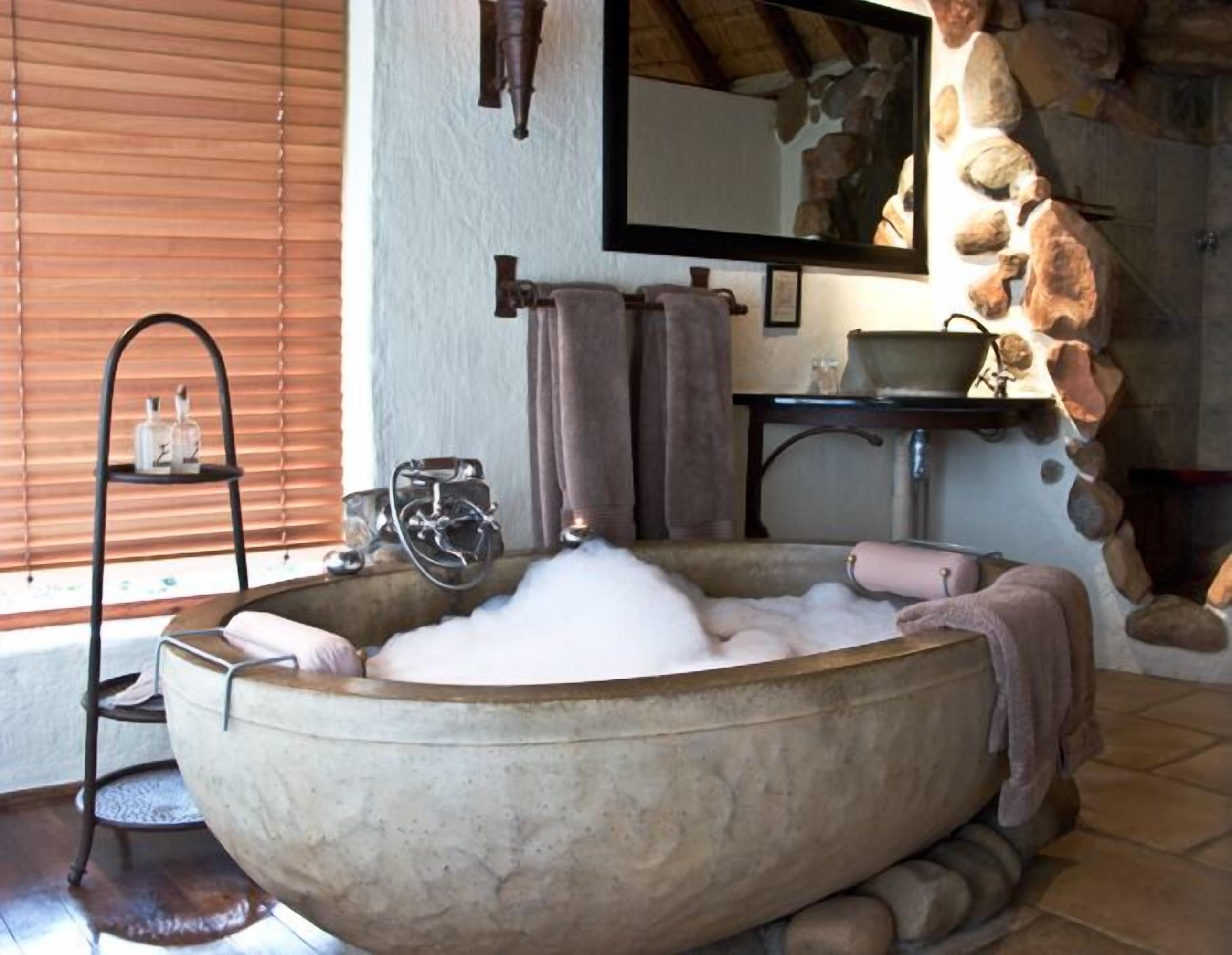 Une grande baignoire en pierre remplie de mousse, une étagère en bois avec des bouteilles, un porte-serviettes et un miroir qui reflète un mur en pierre.