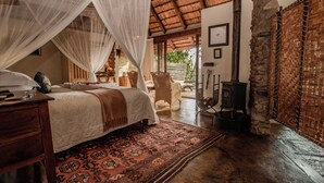 A spacious bedroom with a large bed, a wooden nightstand, a patterned rug, and a stone wall. - Tanamera Lodge (Sabie)