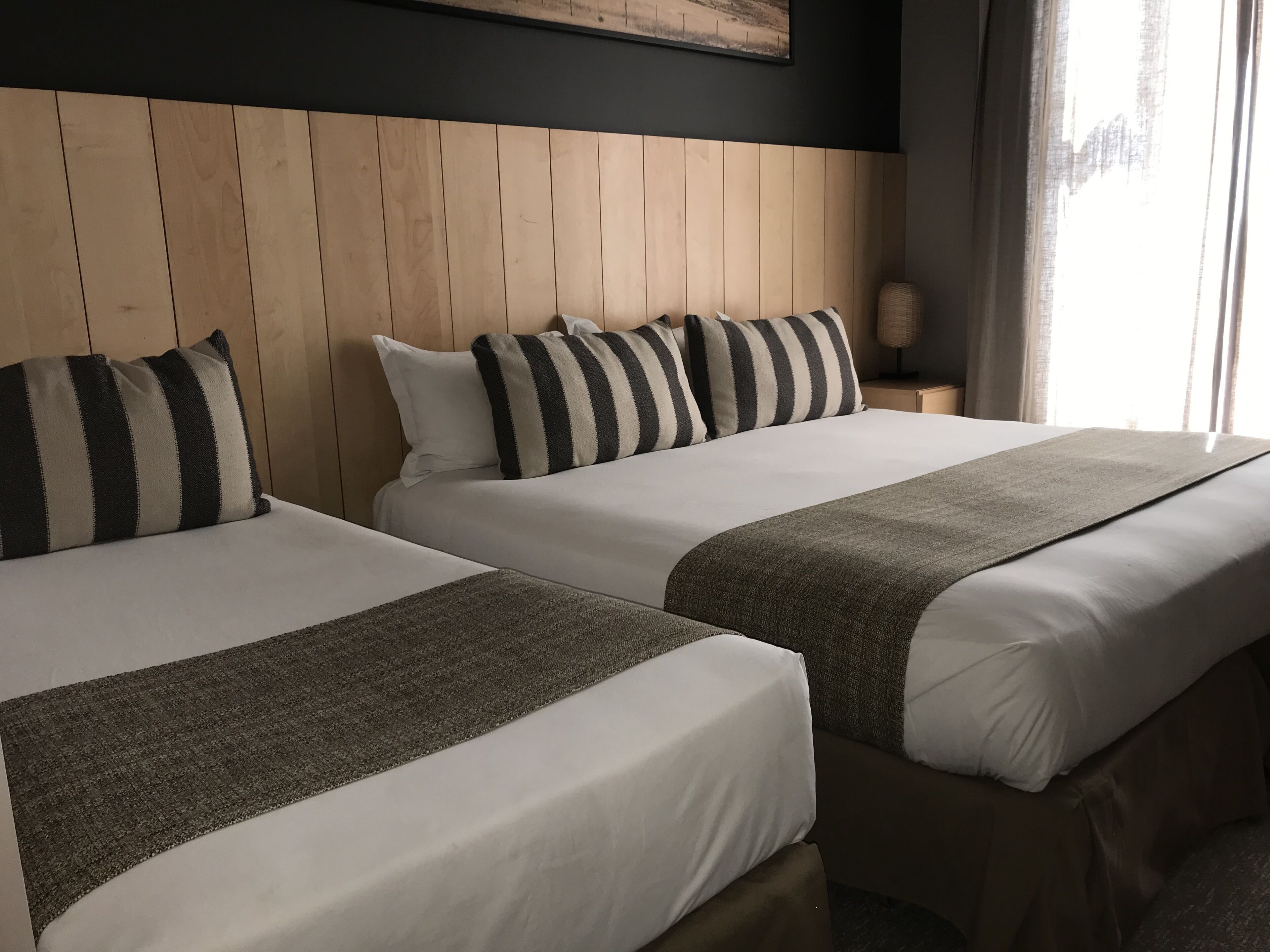 Dos camas con almohadas a rayas y una colcha gris, cabecero de madera, y una mesita de noche con una lámpara.