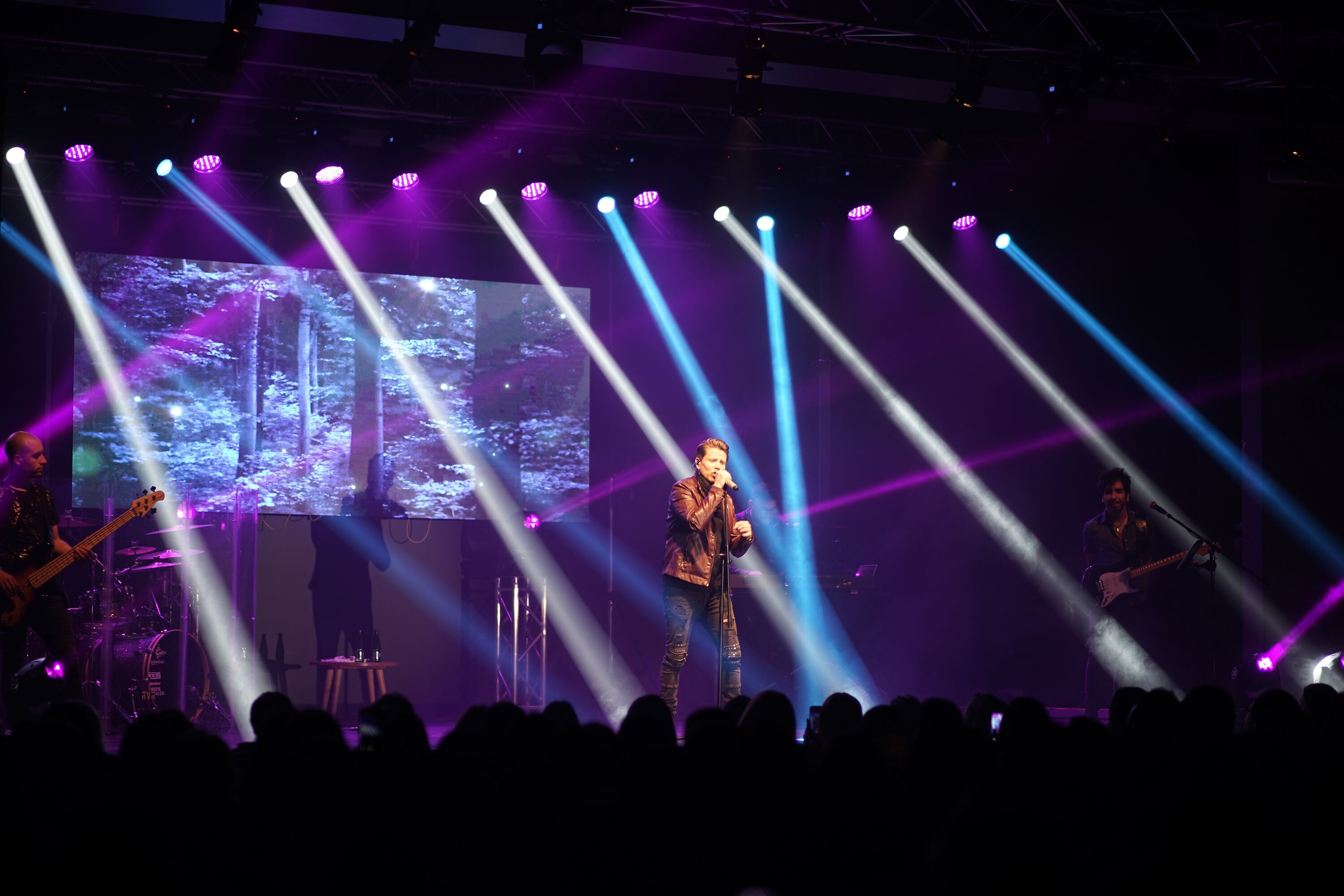 A stage with a band performing, illuminated by stage lights and a large screen displaying a forest scene.