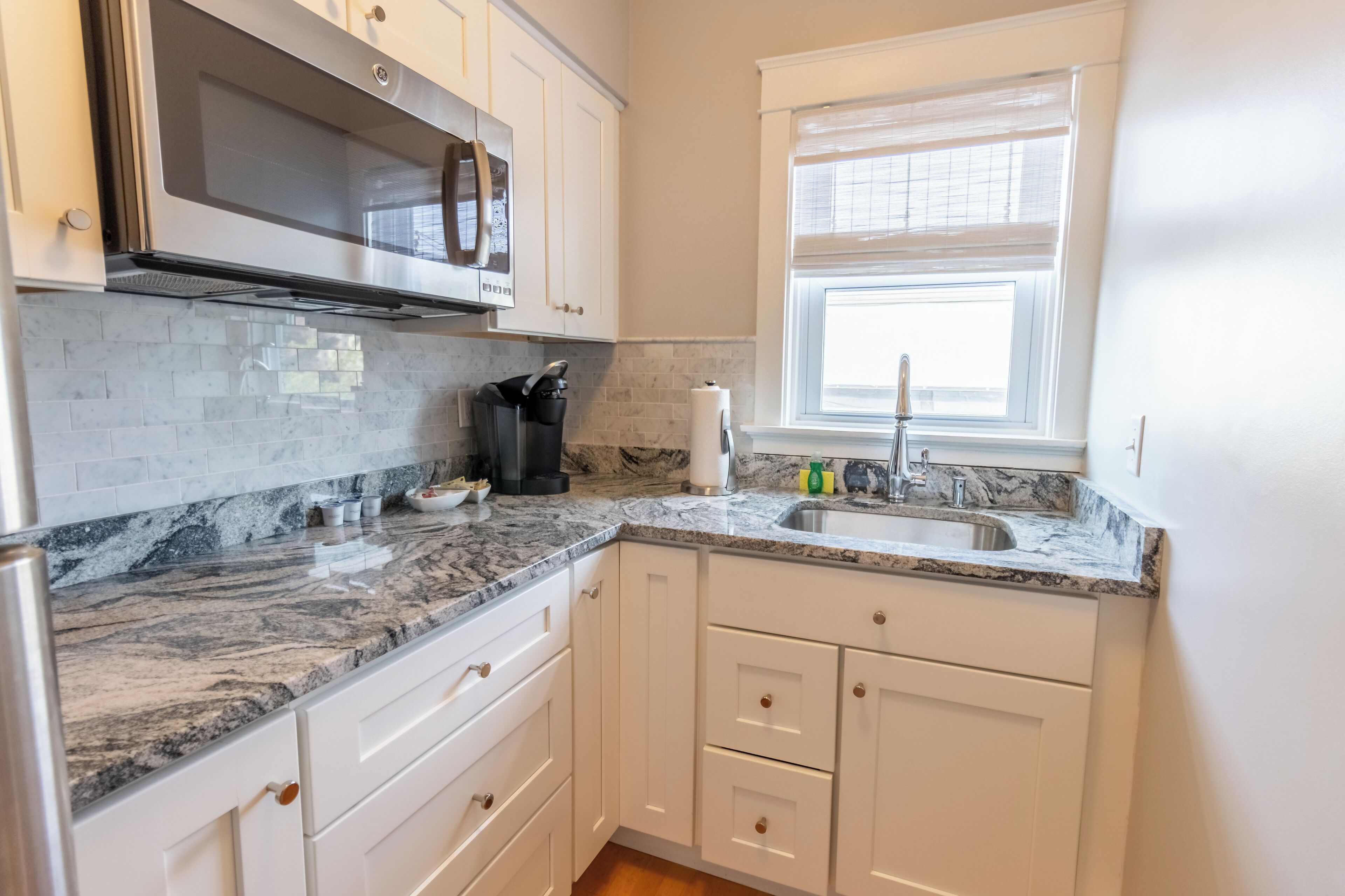 A kitchen with white cabinets, a granite countertop, a stainless steel sink, and a window with blinds.