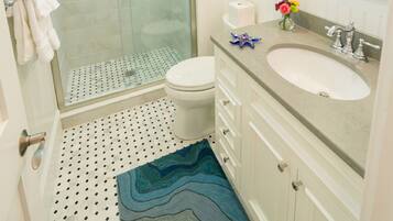A bathroom with a glass-enclosed shower, a white toilet, a sink with a marble countertop, and a patterned rug on the floor.