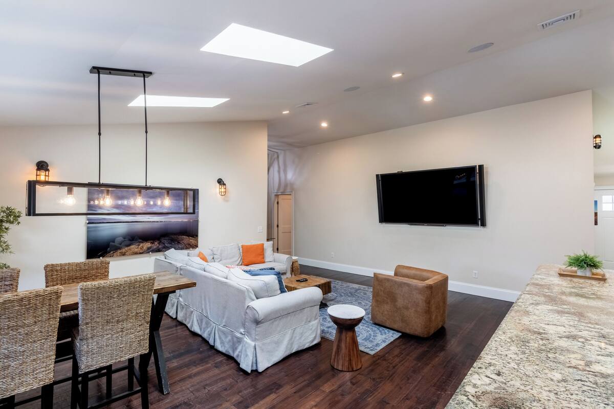 A modern living room with a white sofa, brown armchairs, a dining table with wicker chairs, a flat-screen TV mounted on the wall, and a large window with a view of the outdoors.