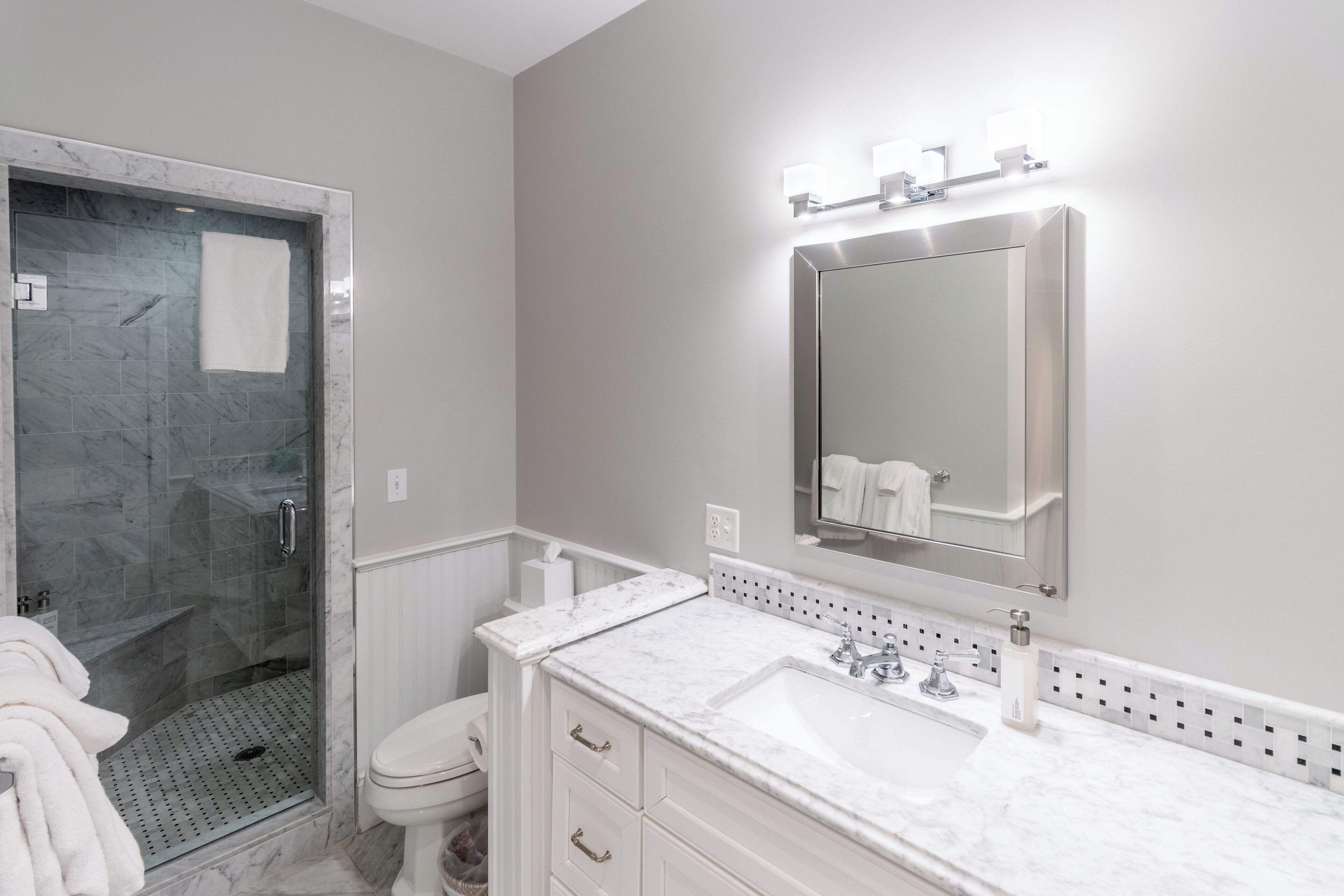 A bathroom with a glass-enclosed shower, a white toilet, a marble sink, and a wall-mounted mirror with lights.