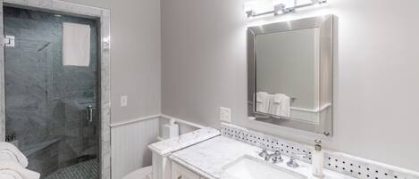 A bathroom with a glass-enclosed shower, a white toilet, a marble sink, and a wall-mounted mirror with lights.