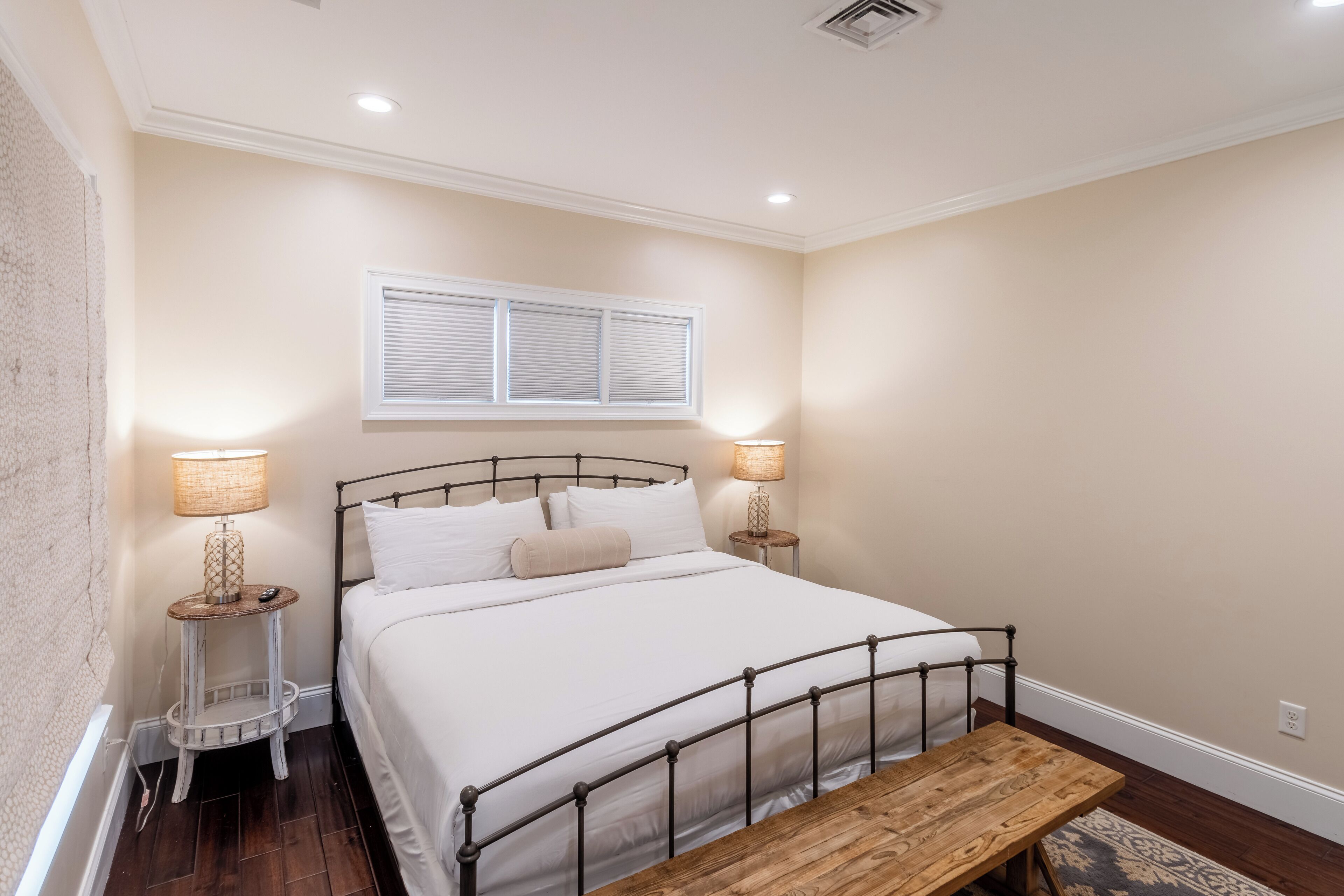 A bedroom with a bed, two bedside lamps, a window with blinds, a small round table, and a bench.