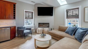 Un salon moderne avec un coin cuisine, un téléviseur à écran plat fixé au-dessus d’une cheminée et une grande fenêtre donnant sur l’extérieur.