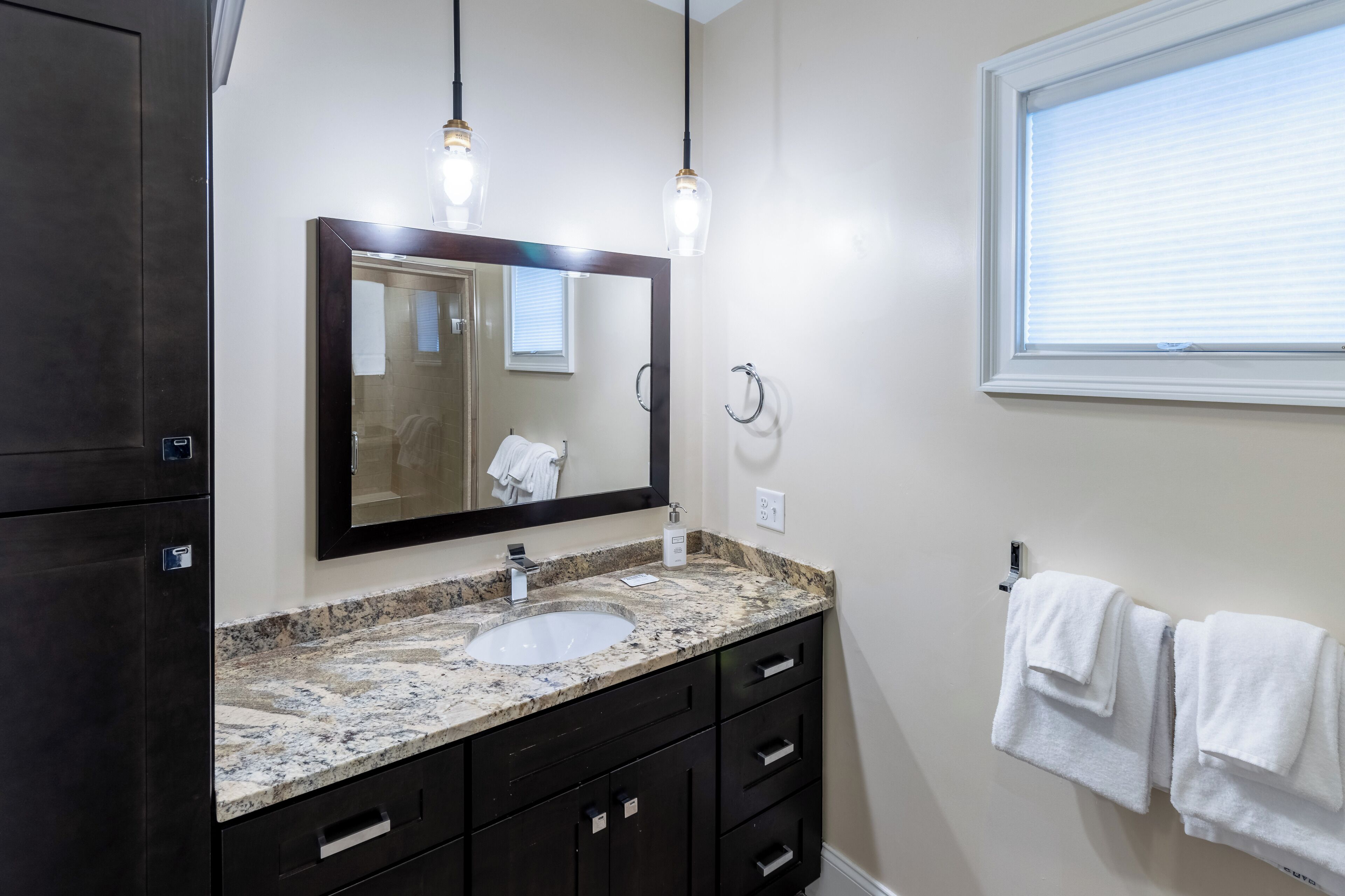 A bathroom with a granite countertop, a sink, dark wooden cabinets, a mirror, and two pendant lights.