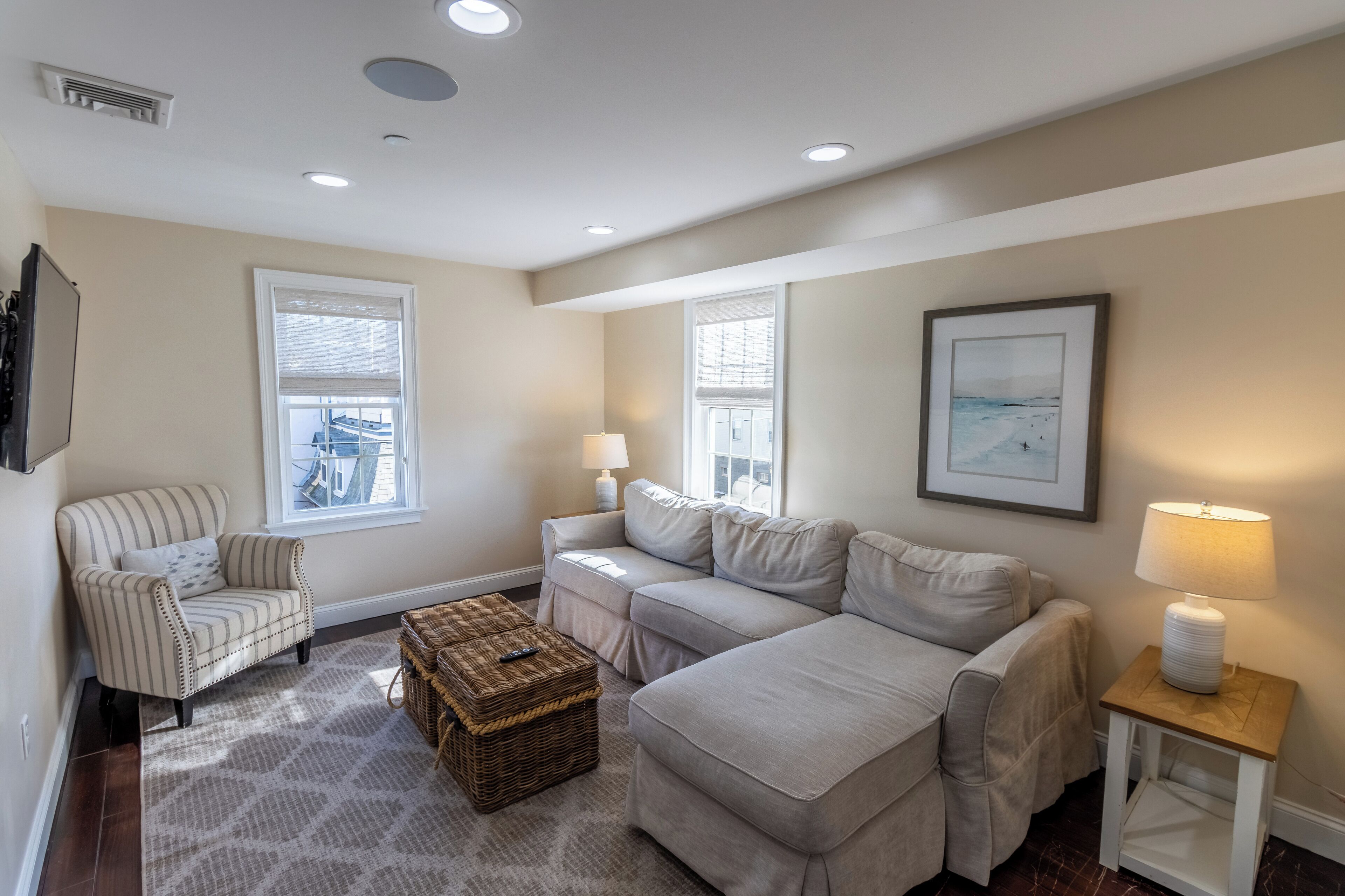 A living room with a striped armchair, a beige sofa, a wicker ottoman, and a side table with a lamp.