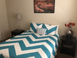 Un dormitorio con cama, mesita de noche, lámpara, florero con flores e imagen enmarcada en la pared.